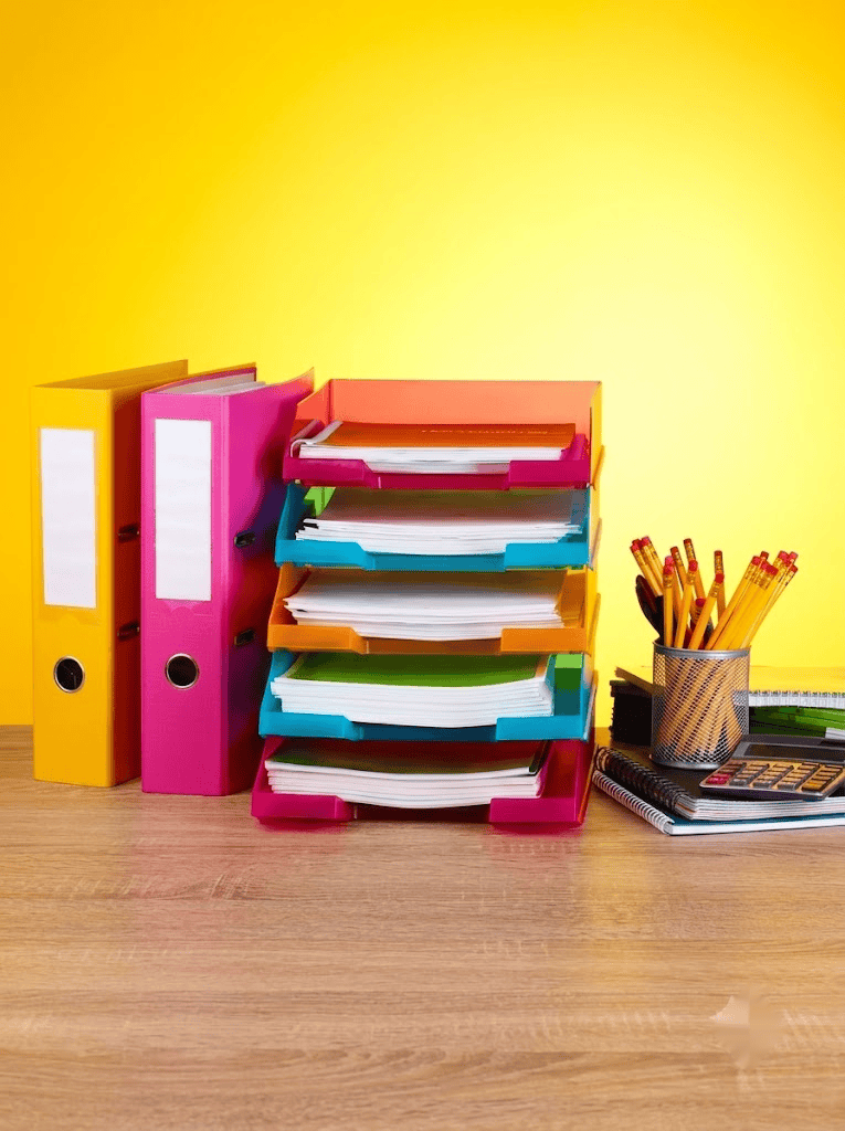 Coloured ring binders, stacked letter trays, pencils and notebooks on a desk